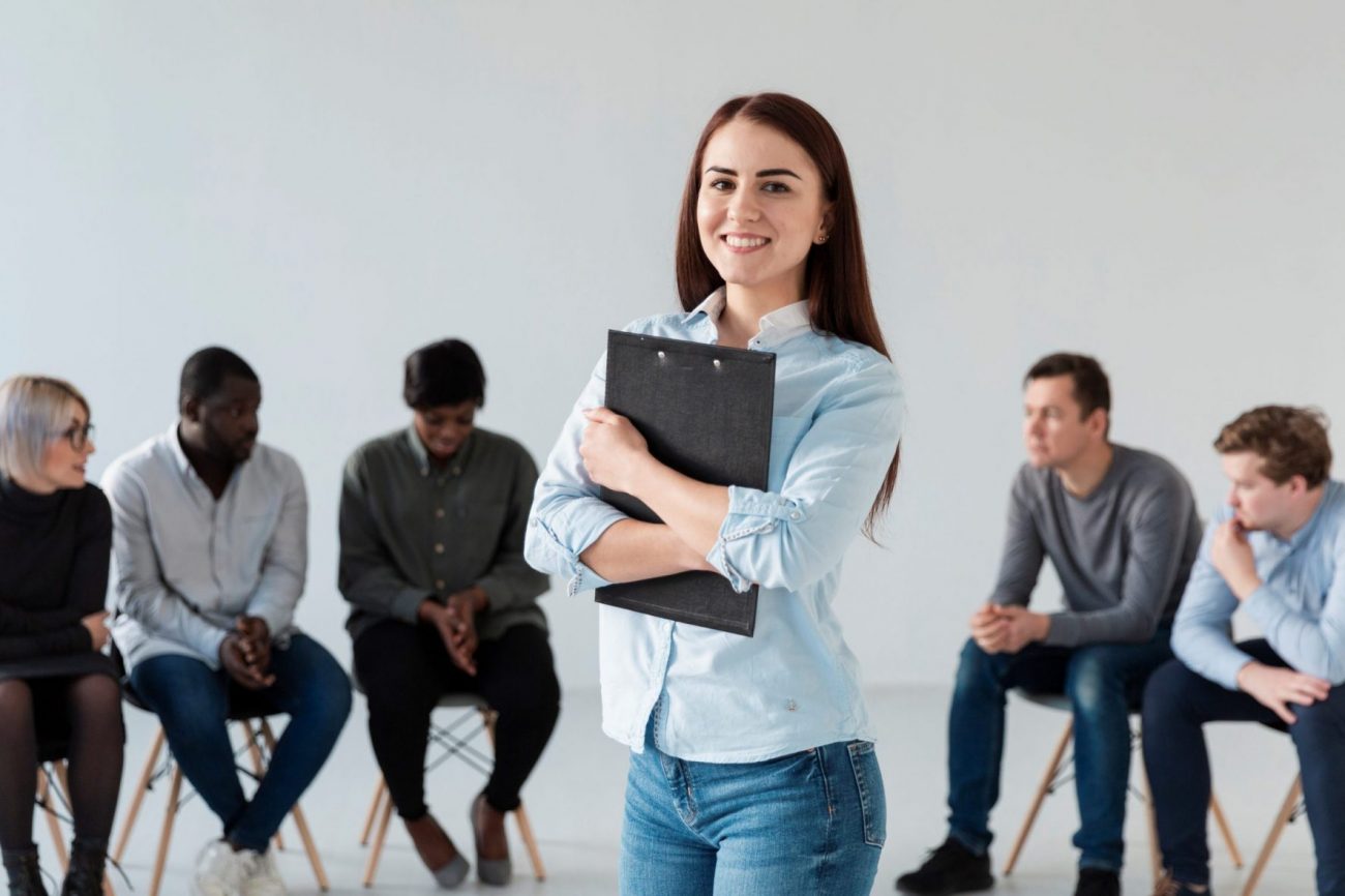 Smiling woman holding clipboard with people sitting in the background illustrates blog "Active vs Passive Candidates: What Is the Difference?"