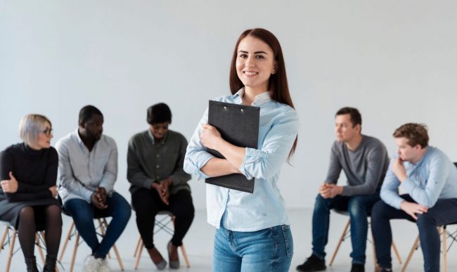Smiling woman holding clipboard with people sitting in the background illustrates blog "Active vs Passive Candidates: What Is the Difference?"