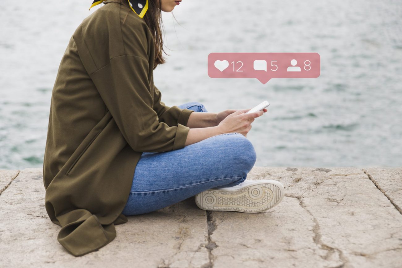 social-media-fp Woman wearing jeans and a green jacket sitting on the floor holding smartphone with social media icons hovering above it.